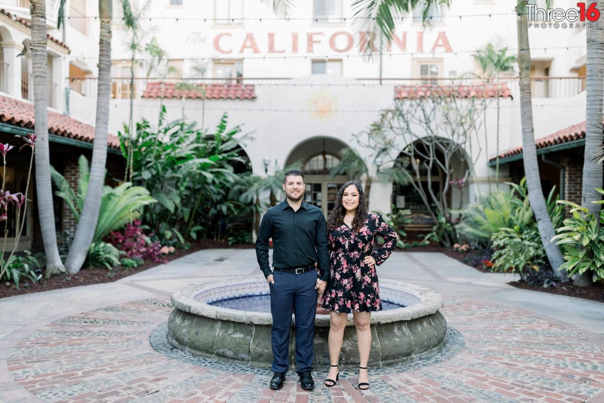 Engaged couple stand side by side in front of the Villa Del Sol water fountain