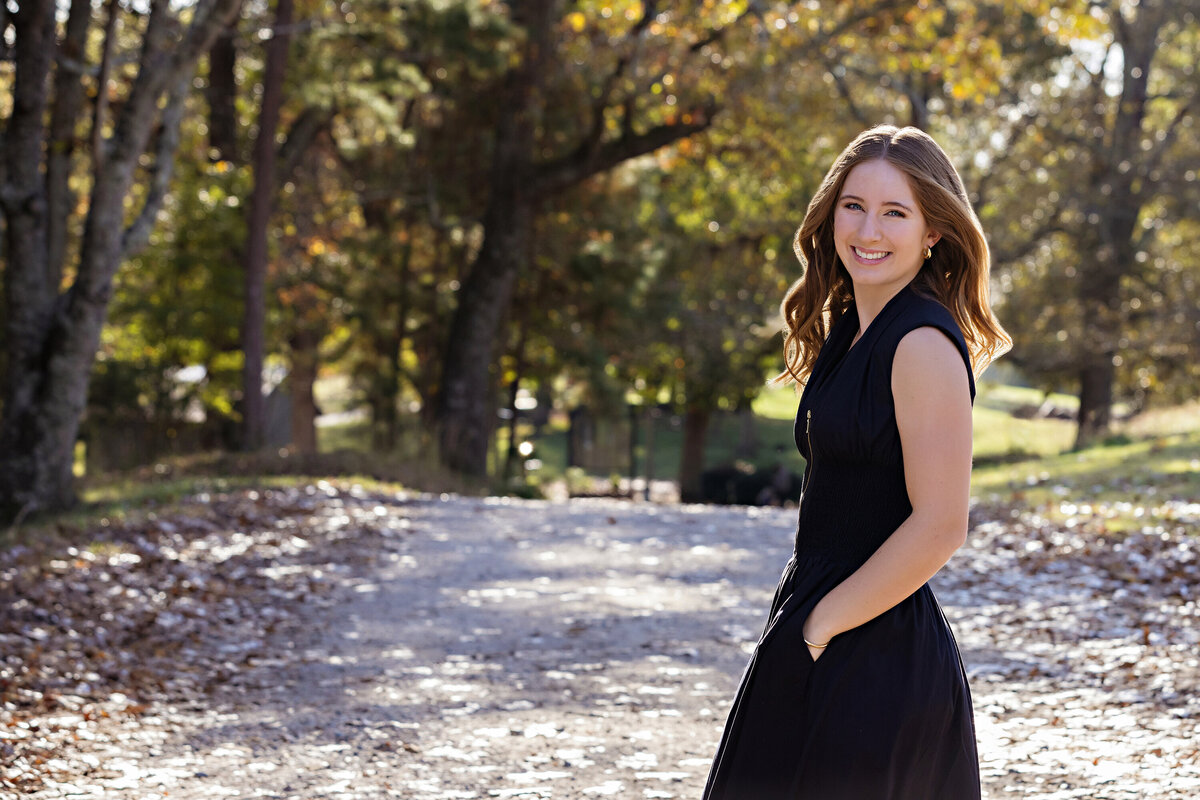 High school senior in black dress with gravel path and glowing yellow trees in Charlotte area