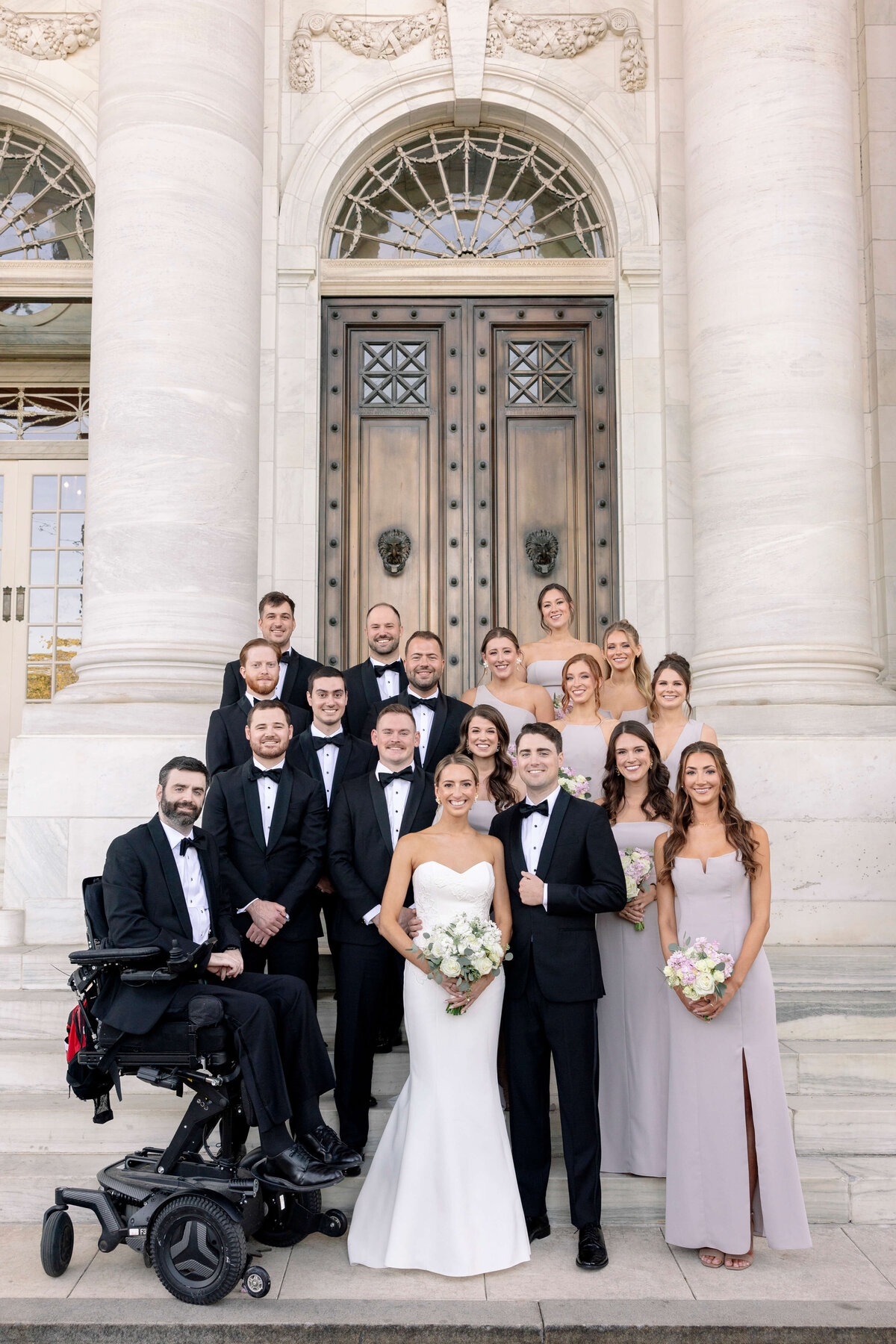 A bride and groom stand with their wedding party on stone steps in front of a large building with ornate doors. The group includes men in tuxedos and women in light dresses, with one man in a wheelchair. Everyone is smiling and holding flowers.