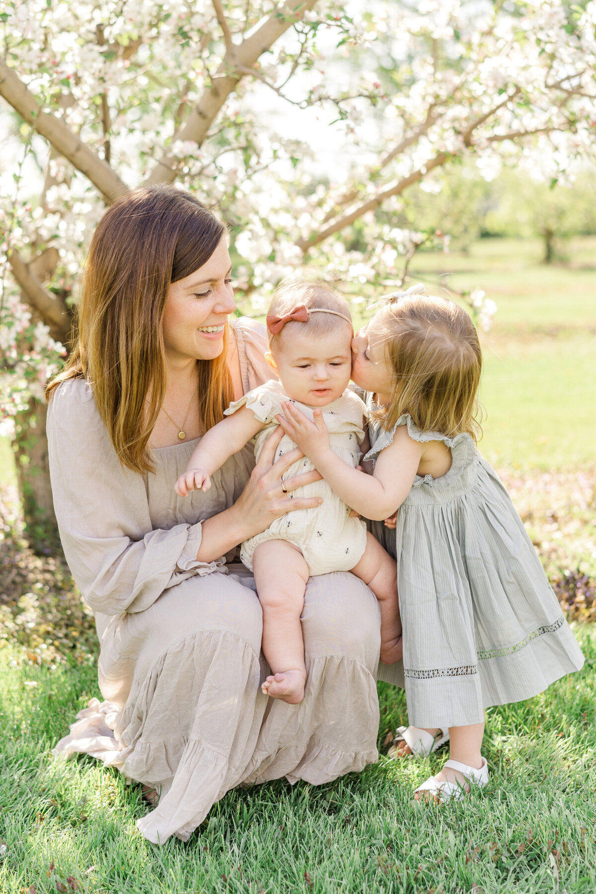 mother and two daughters in the grass together while the older sibling kisses the younger one