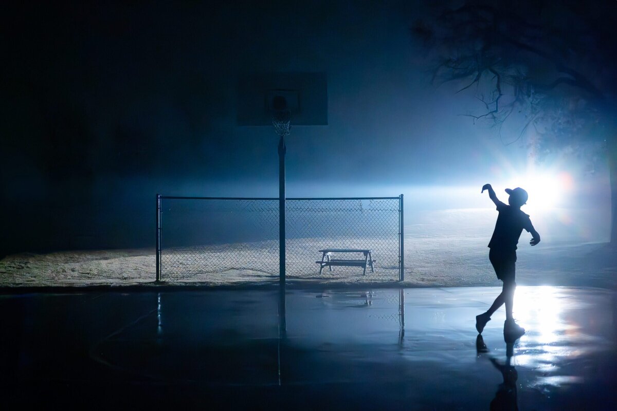 A car's headlights illuminate a young basketball player as he throws the ball at dusk.