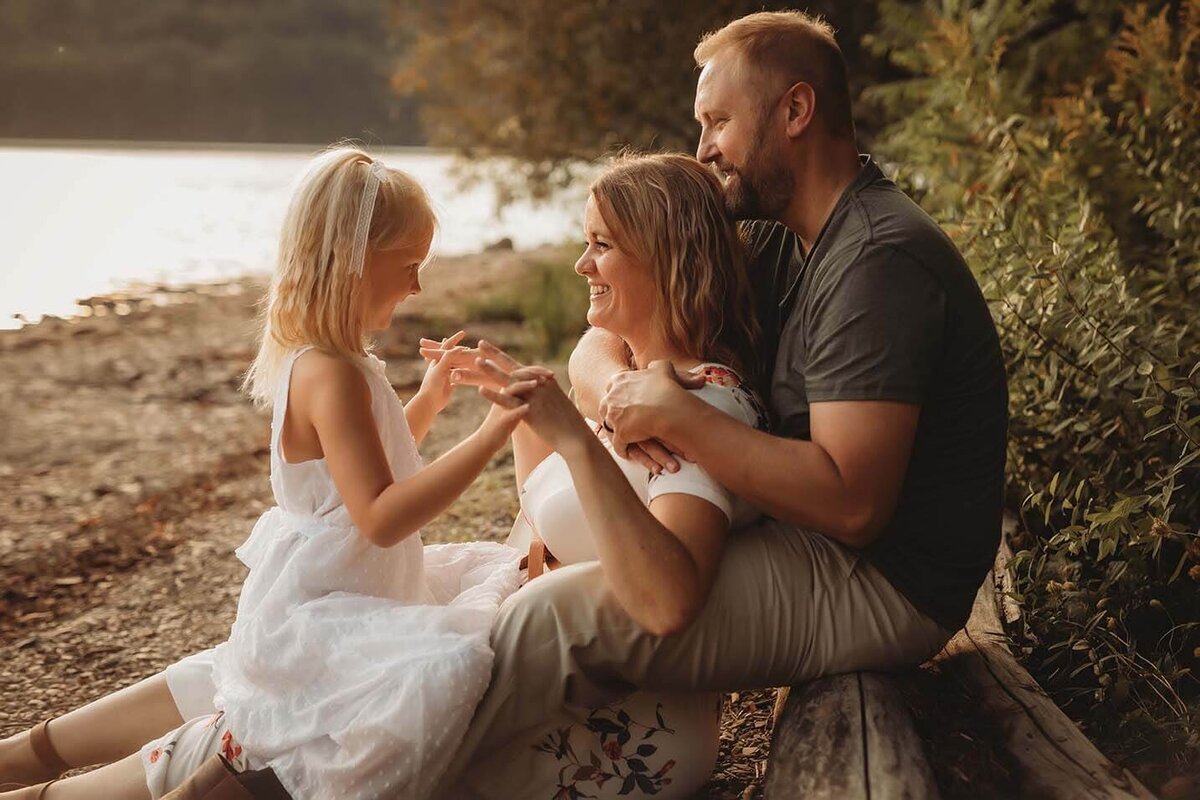 a dad sits on a log while his wife sits in his lap. their daughter sits on the moms lap  while they play and hold hands in rochester ny