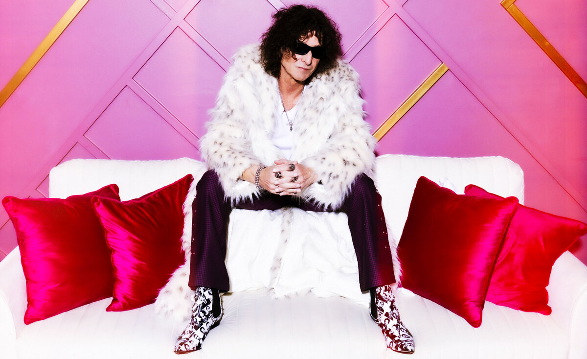Male musician portrait Venrez wearing white fur coat hands together sitting on white sofa with pink background