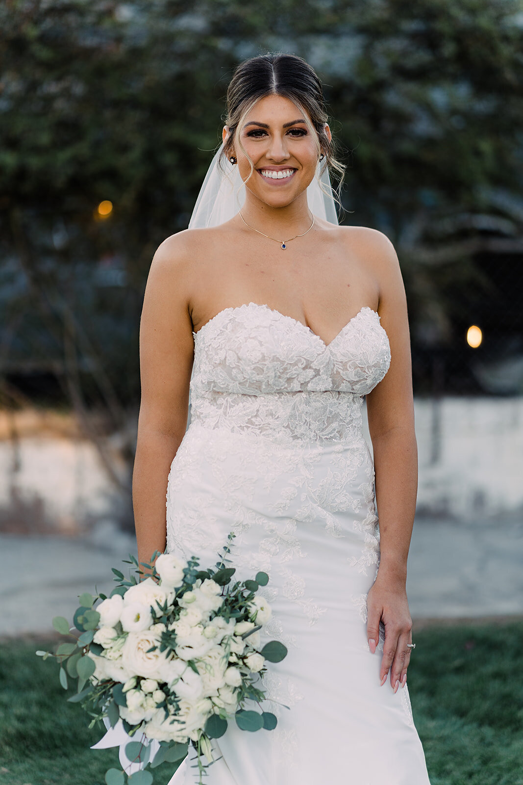 The bride, in a fitted, lace-embellished strapless gown, smiles directly at the camera, holding a bouquet of white flowers and greenery.