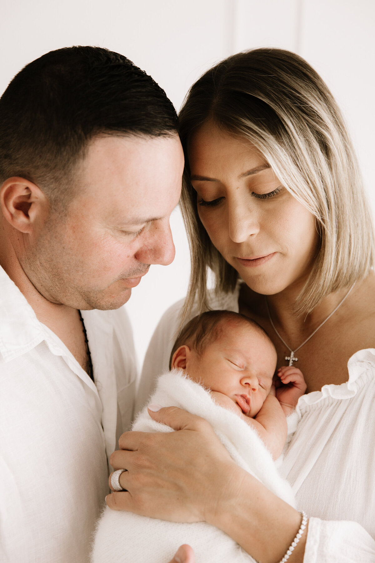 Close-up of a mother and father lovingly gazing at their swaddled newborn baby in a soft, white Southwest Sydney studio setting.