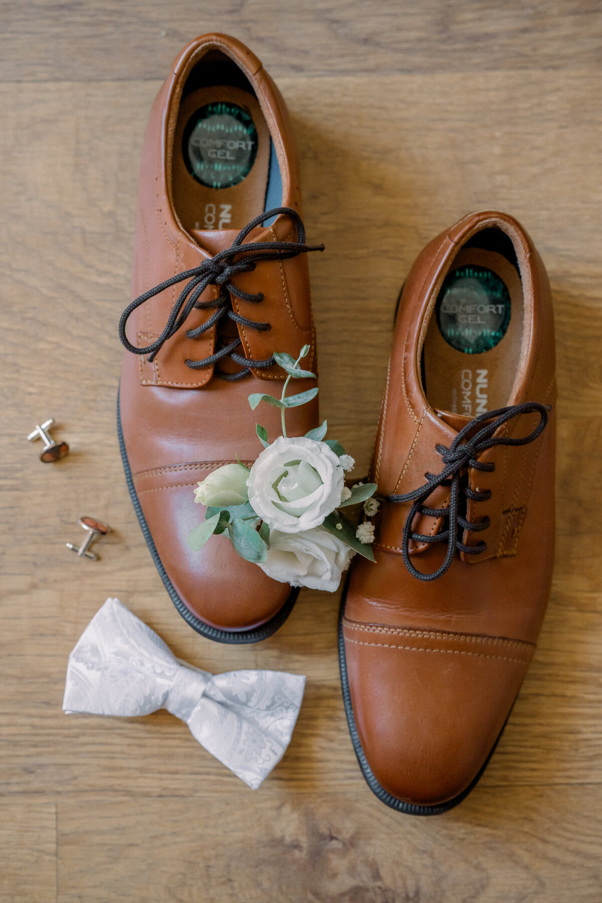 Weddings in Spain | Brown dress shoes, custom cufflinks, a white bow tie, and a white floral boutonniere lay on the hardwood floor of a Spanish home.