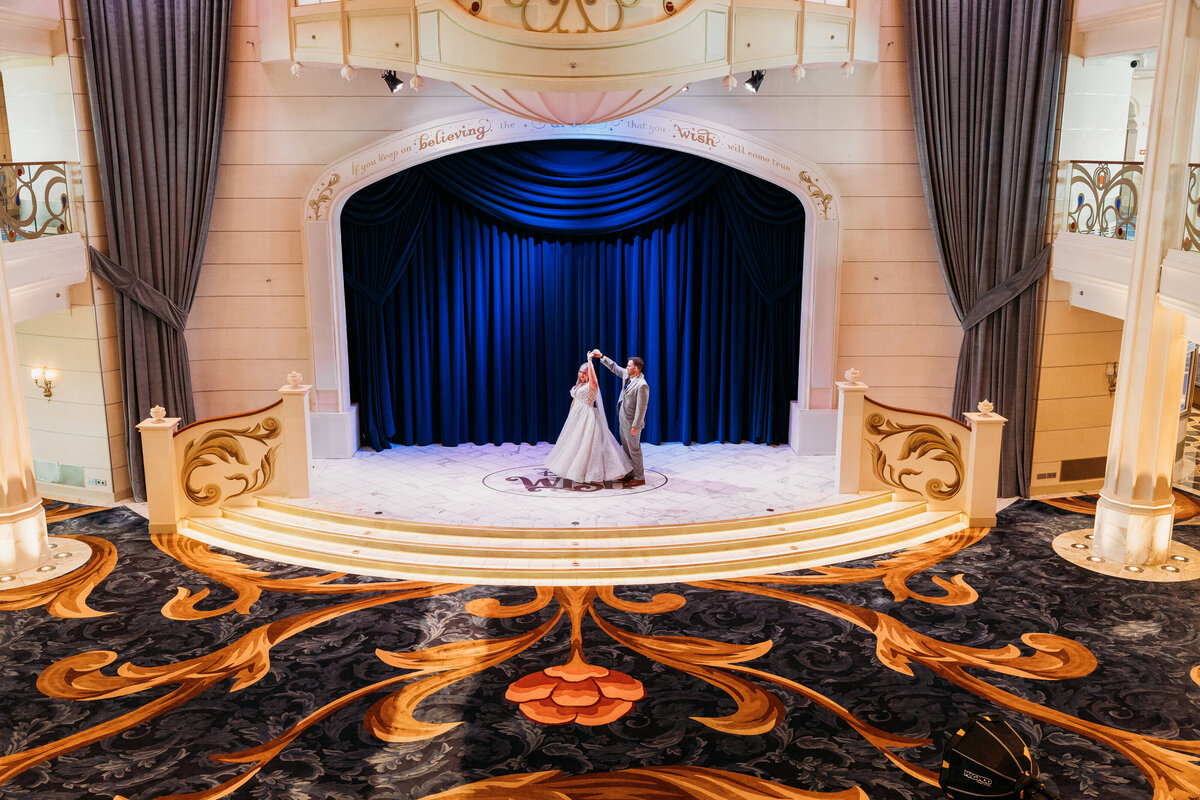 Bride and groom twirl on the stage inside the Disney Wish Grand Hall beneath a quote about wishes and magical fairy tale dreams.