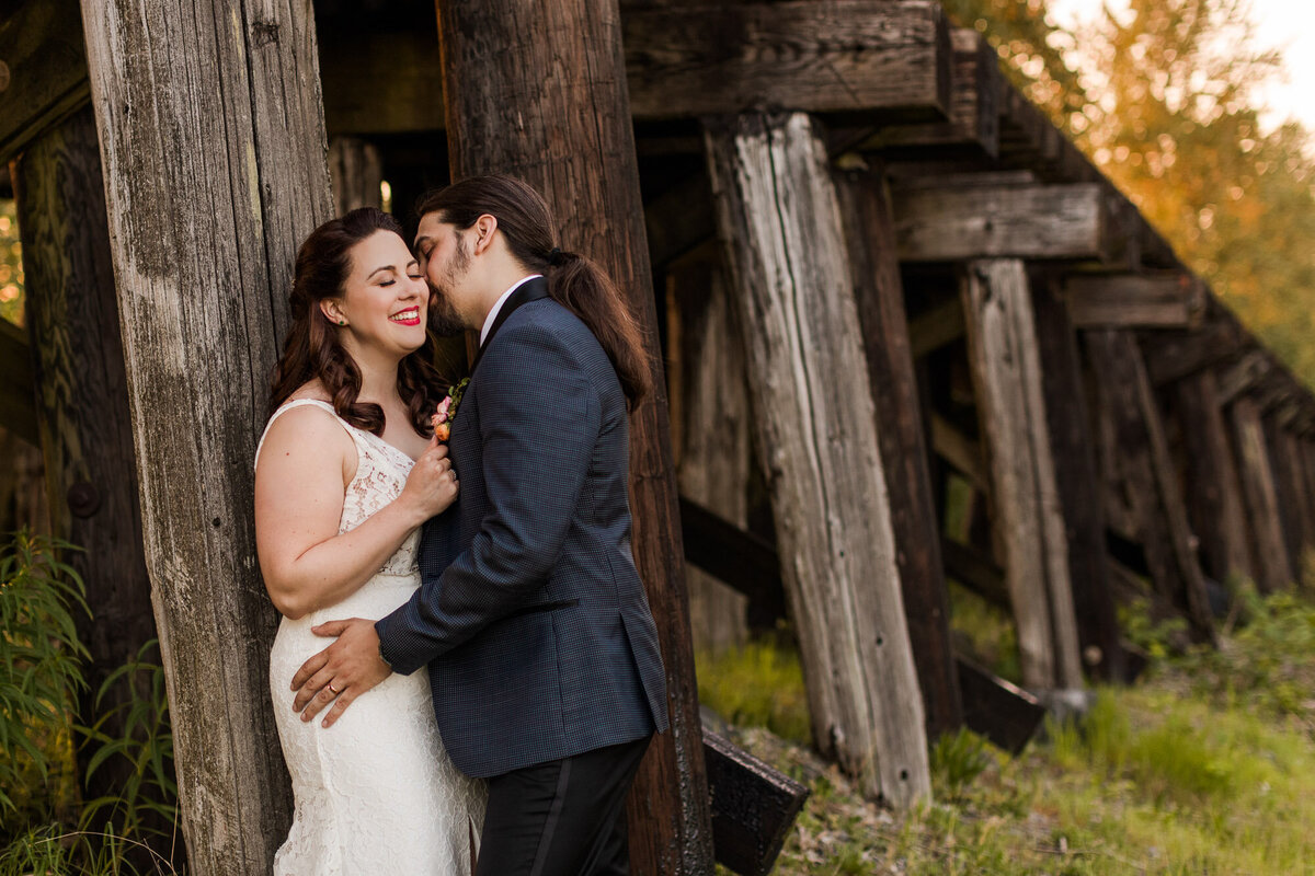 Bride-and-groom-kiss-underneath-a-railroad-trestle-during-golden-hour-on-their-wedding-day-at-venue-Hidden-Meadows-in-Snohomish-photo-by-Joanna-Monger-Photography