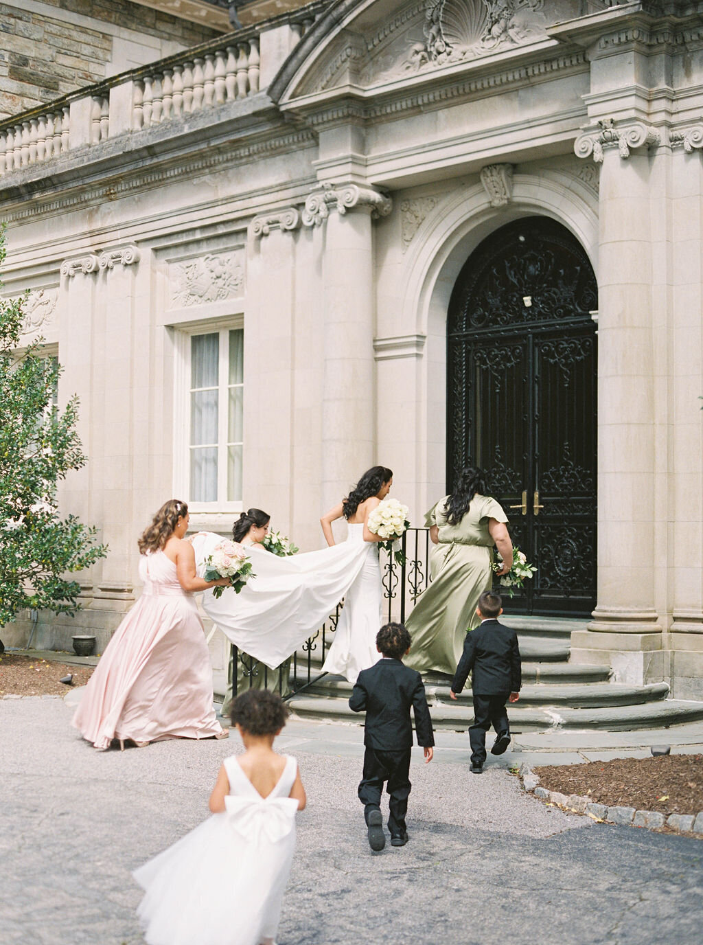 Wedding Photographer RI | Bridesmaids in flowing dresses and a bride with a long train ascend stairs towards an ornate building entrance, followed by young children.