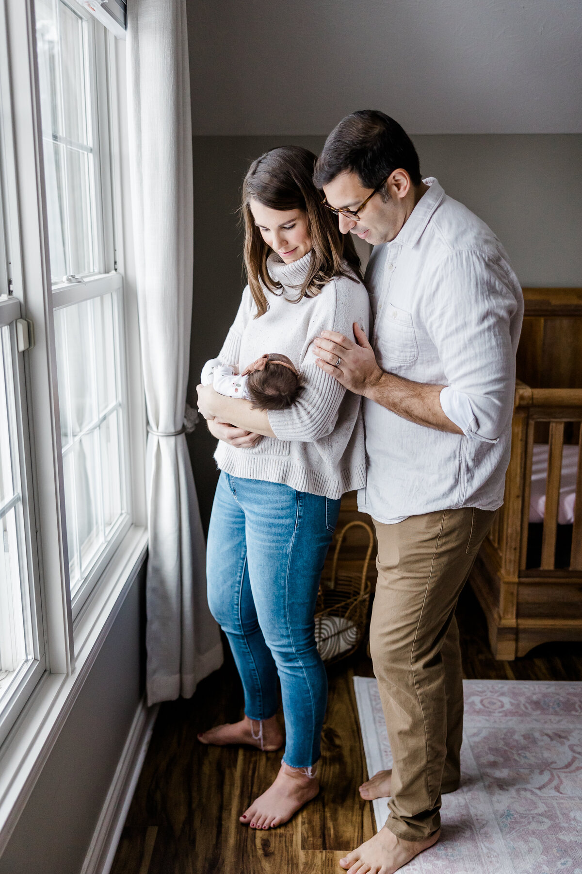 mom and dad standing in front of window in nursery snuggling newborn