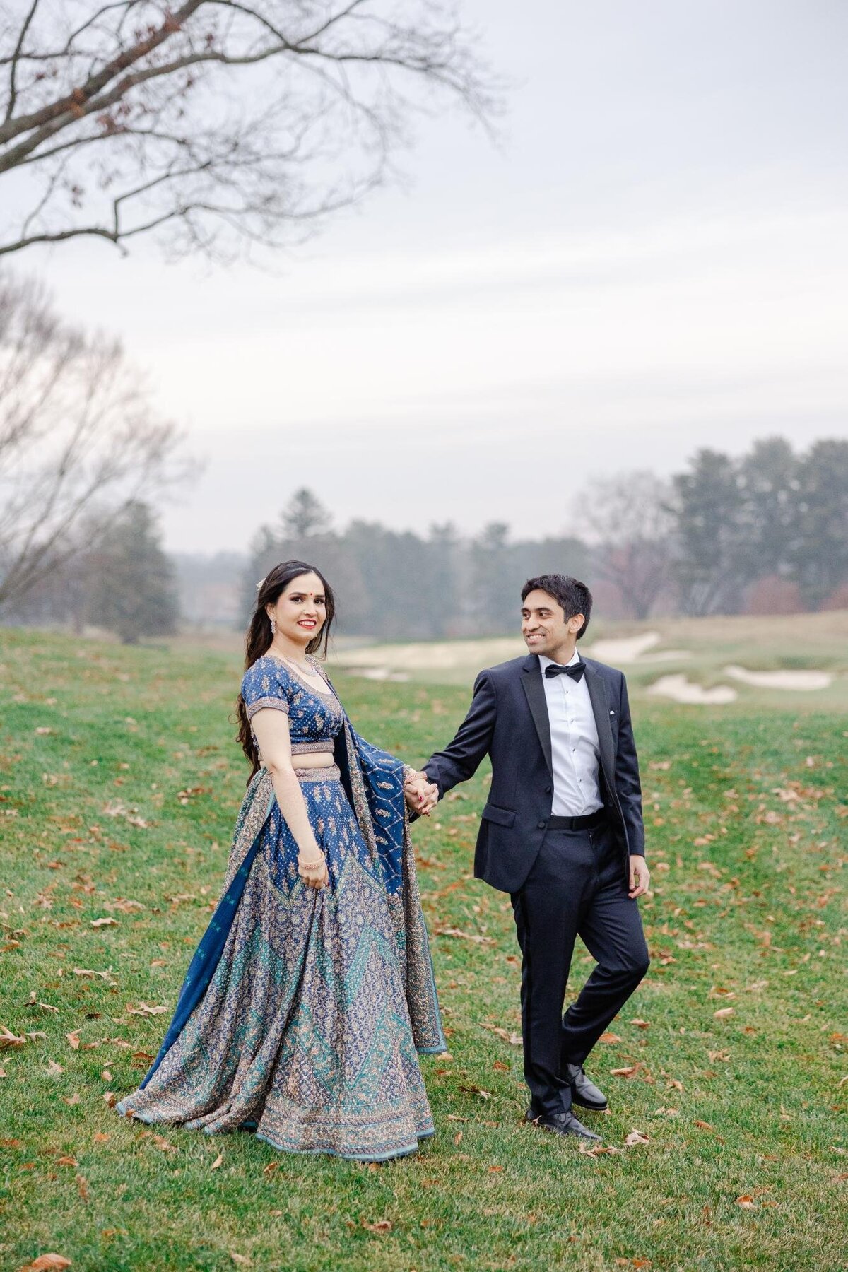 A couple in formal attire, the woman in a flowing blue dress and the man in a black tuxedo, walking hand in hand on a grassy field with a foggy background.