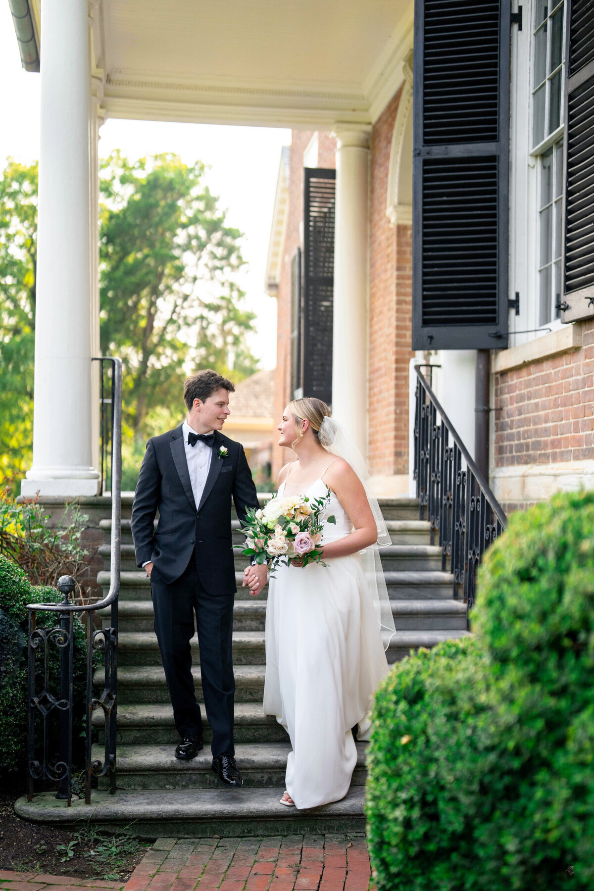 A bride and groom, dressed in a white gown and black suit respectively, smile at each other while walking down steps outside a brick building. The bride holds a bouquet of flowers. Lush greenery surrounds them on a sunny day.