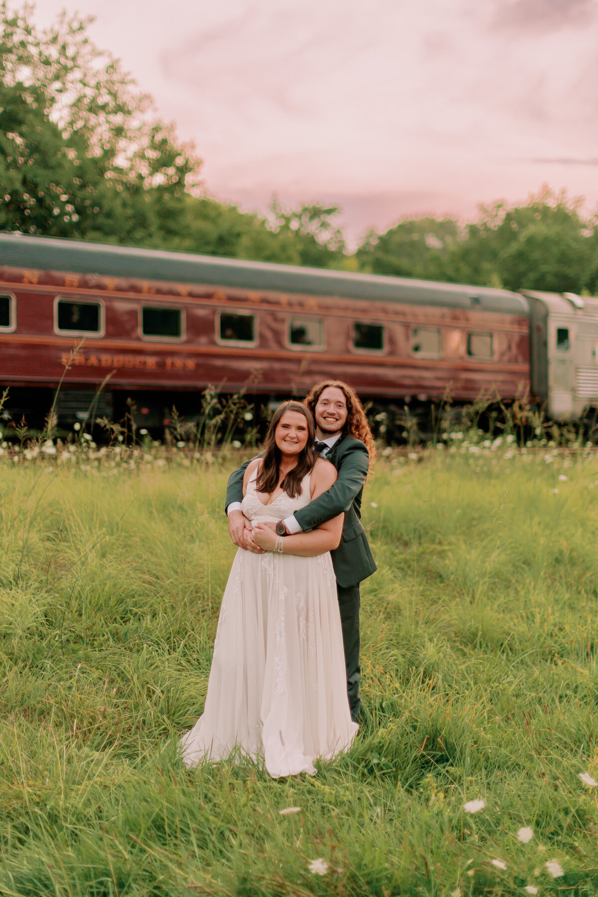 A person standing behind their partner smiling in front of an old train 