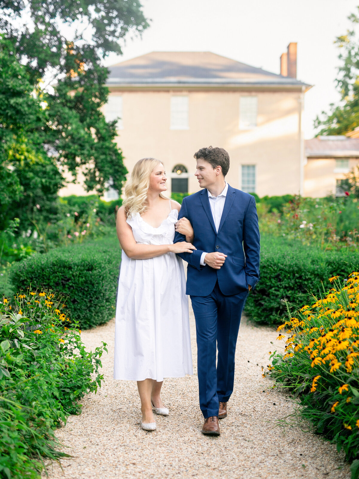 A couple walks arm-in-arm on a gravel path in a garden. The woman wears a white dress, and the man is in a blue suit. They are smiling at each other, surrounded by green hedges and yellow flowers, with a house in the background.