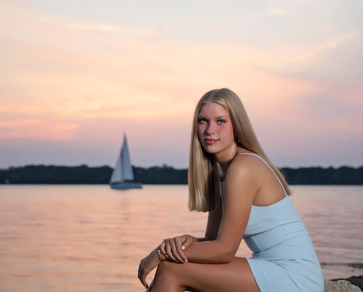 Senior photos of a girl sitting at the Erie Yacht Club at sunset with a sailboat in the distance