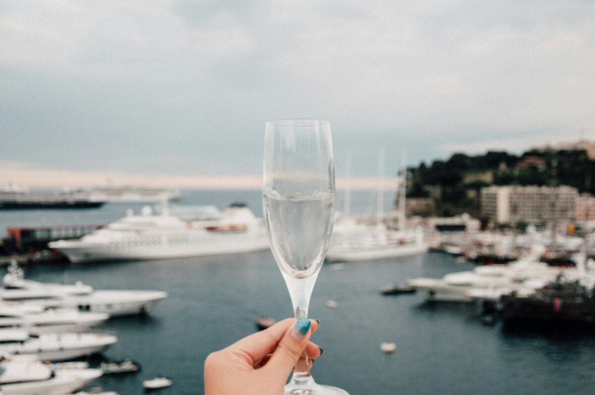 A hand with painted nails holds a champagne glass in the foreground, with a marina full of yachts and boats in the background