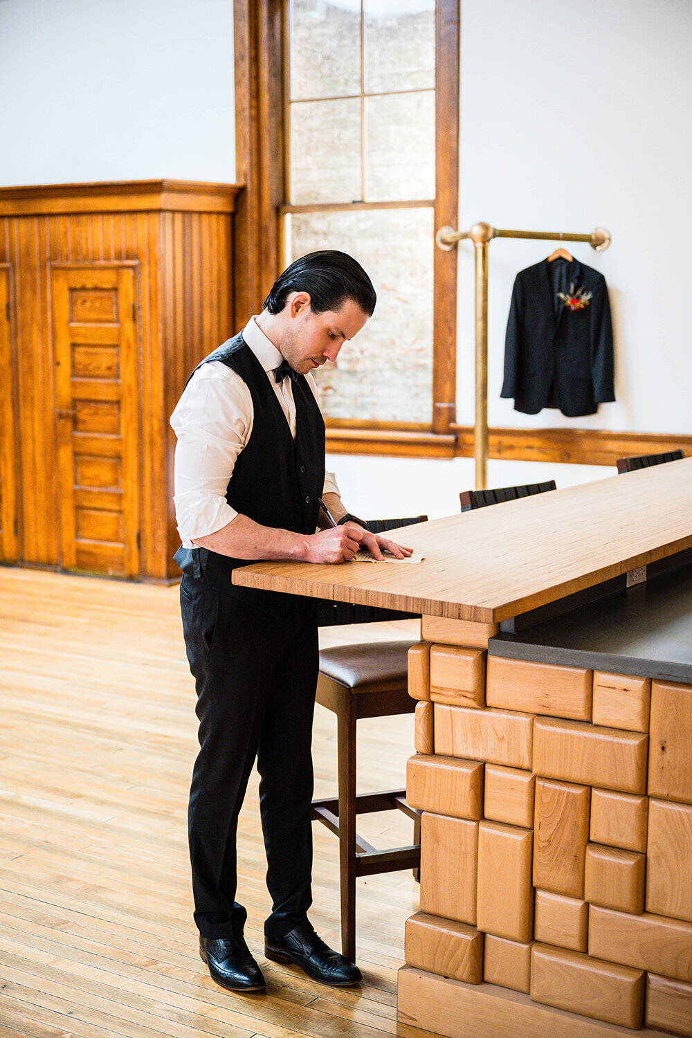 A groom stands at the bar in the Bunkroom at Fire Station One to write his vows for his elopement ceremony with his suit jacket hanging in the distance.