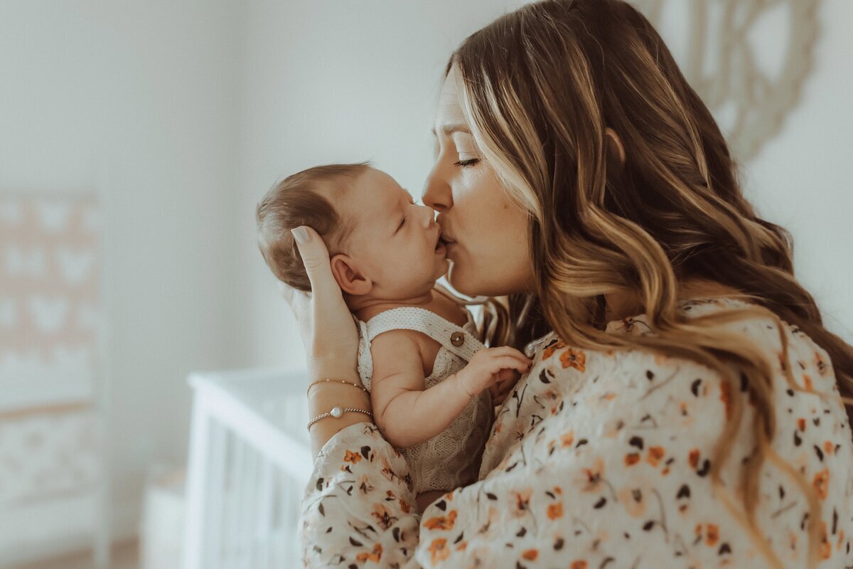 A woman with long hair kisses a baby on the cheek while holding them in a softly lit room.