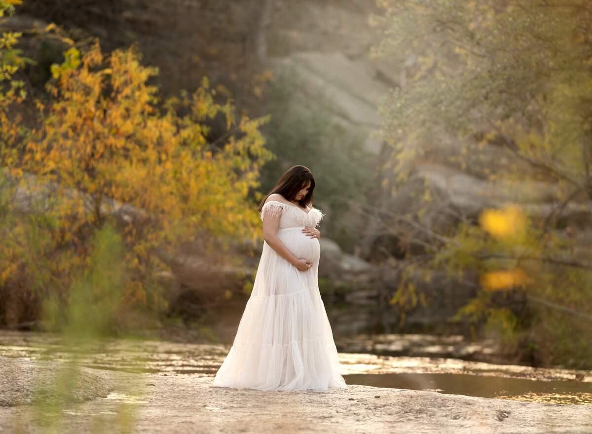Woman in soft white dress cradling her belly by the water with golden fall leaves at Bull Creek District Park