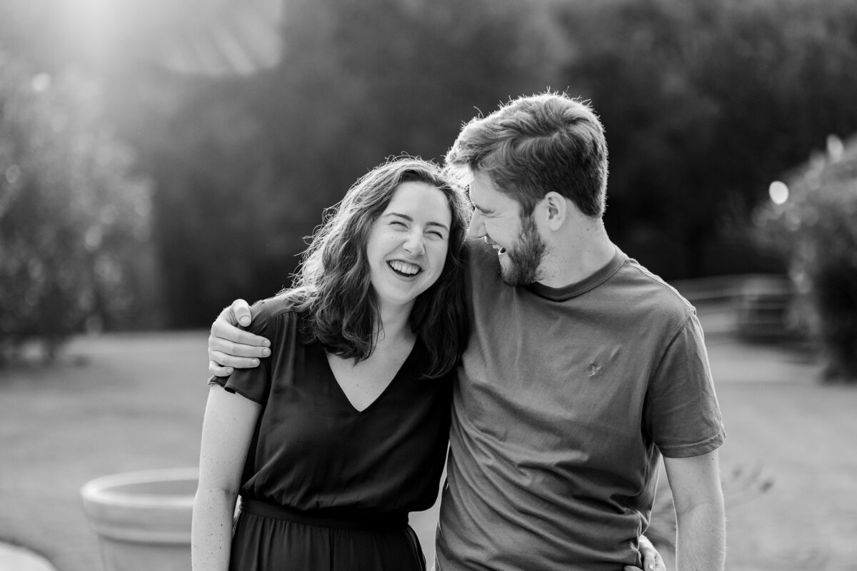 A black and white candid photo of a couple laughing and holding each other close during their engagement session in DFW, Texas. The woman on the left is wearing a short sleeve dress while the man on the right is wearing a t-shirt. They are backed by many trees in the background.