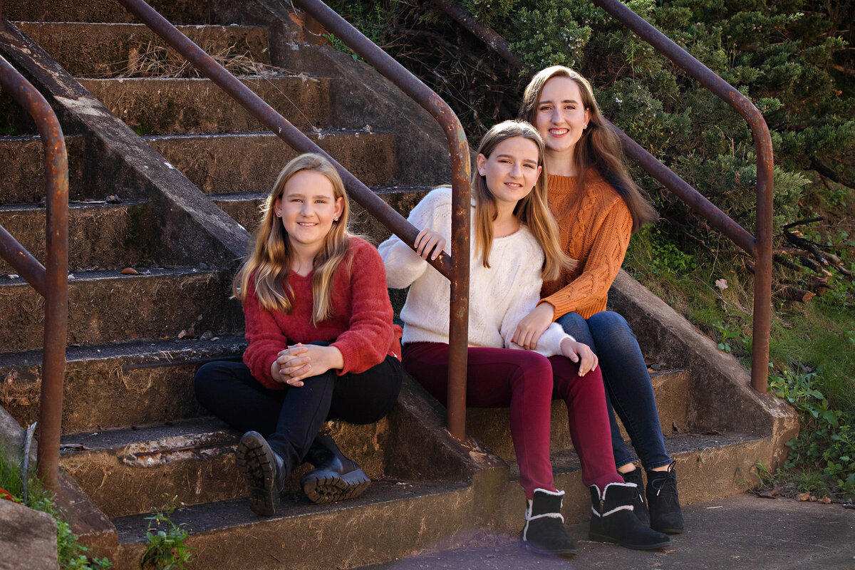 Smiling siblings in autumn colors seated on concrete staircase surrounded by greenery at Camp North End Charlotte NC