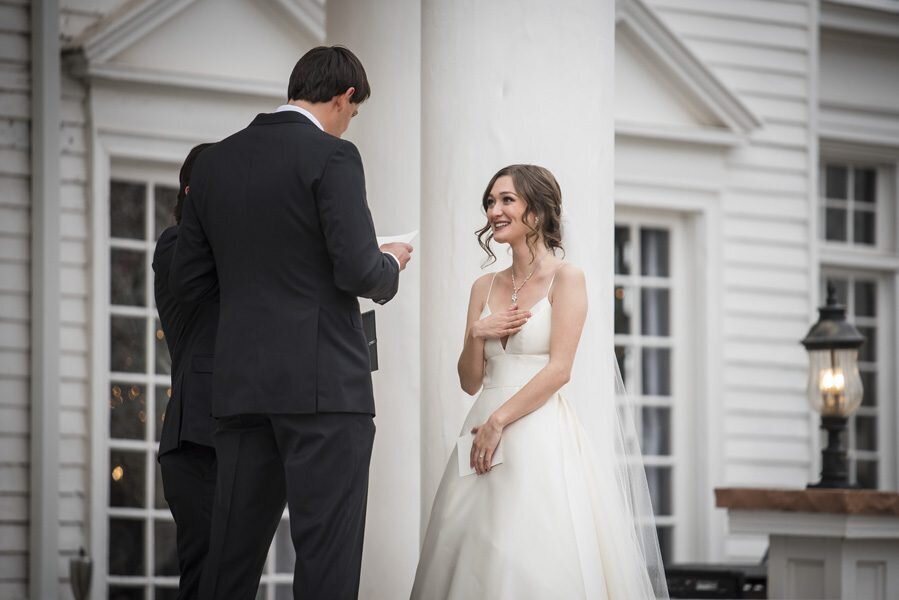 A bride places her hand over her heart as her groom reads his vows to her on the steps of The Manor House in Littleton, Colorado.