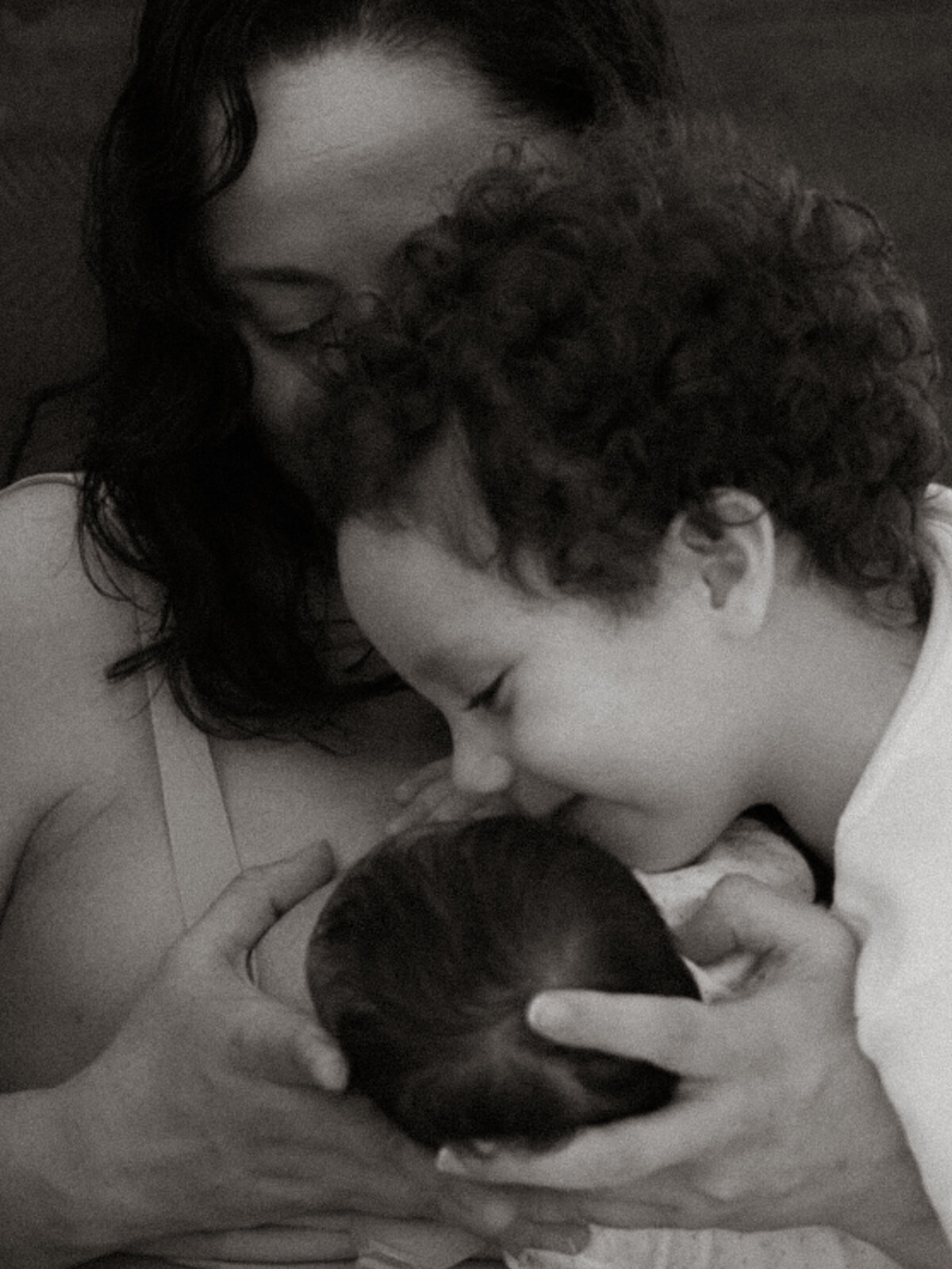 A young brother gently kissing the head of his newborn sibling while being held by their mother, in a soft black-and-white portrait.