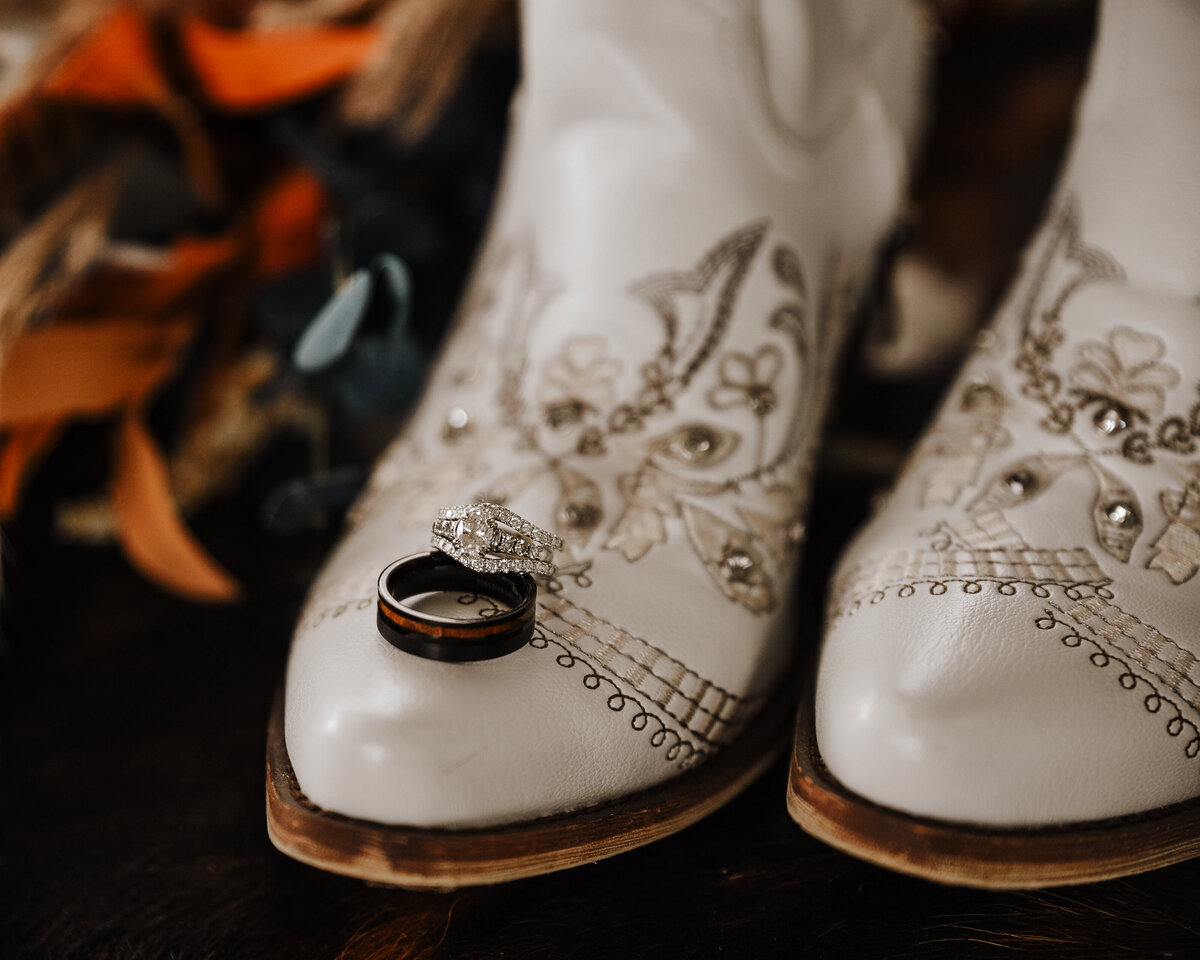 Wedding elegance with a country twist – a pair of white embroidered cowboy boots showcasing a set of wedding rings taken by jen Jarmuzek photography a Minneapolis wedding photographer