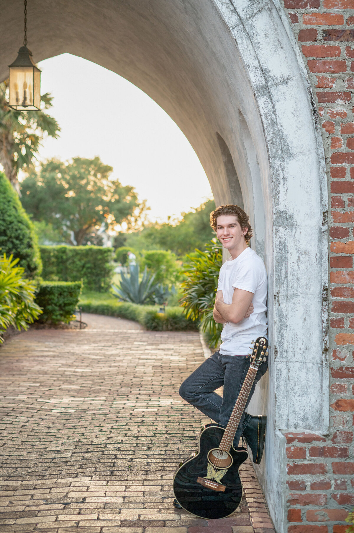 High school senior boy leaning against a stone archway with guitar in front of him smiling.