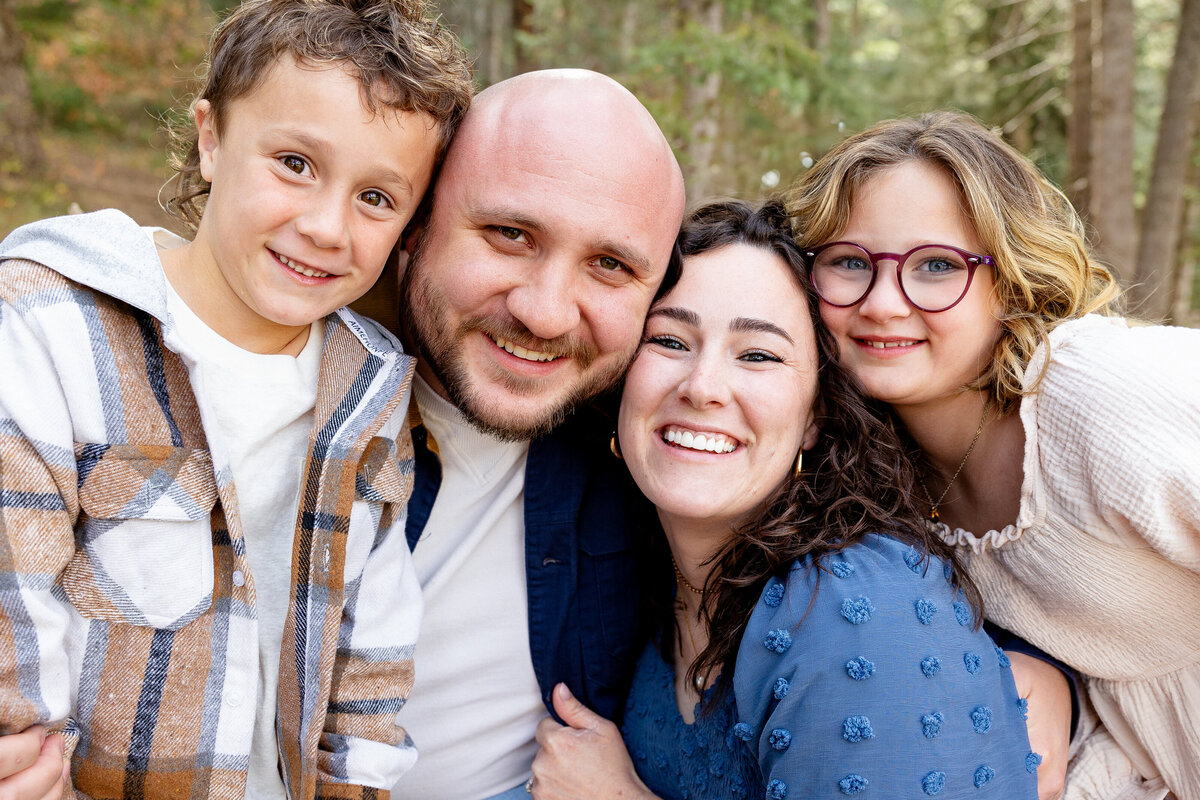 A close-up shot of a joyful family of four, embracing closely and smiling brightly. The father, mother, and their two children share a moment of happiness in a beautiful wooded setting. Taken by Cassie Lee Photography