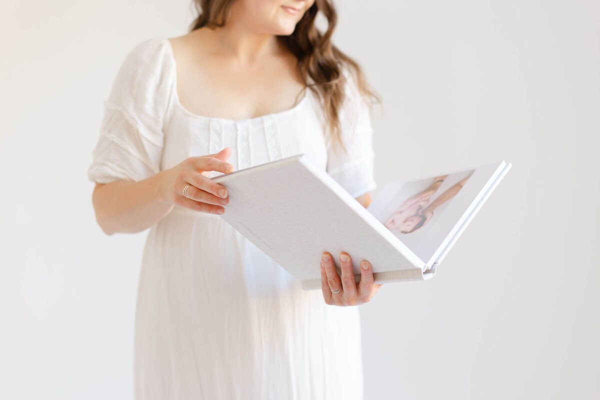 Portrait of a woman in a white dress holding a linen newborn album. Her face is not showing in the photograph.