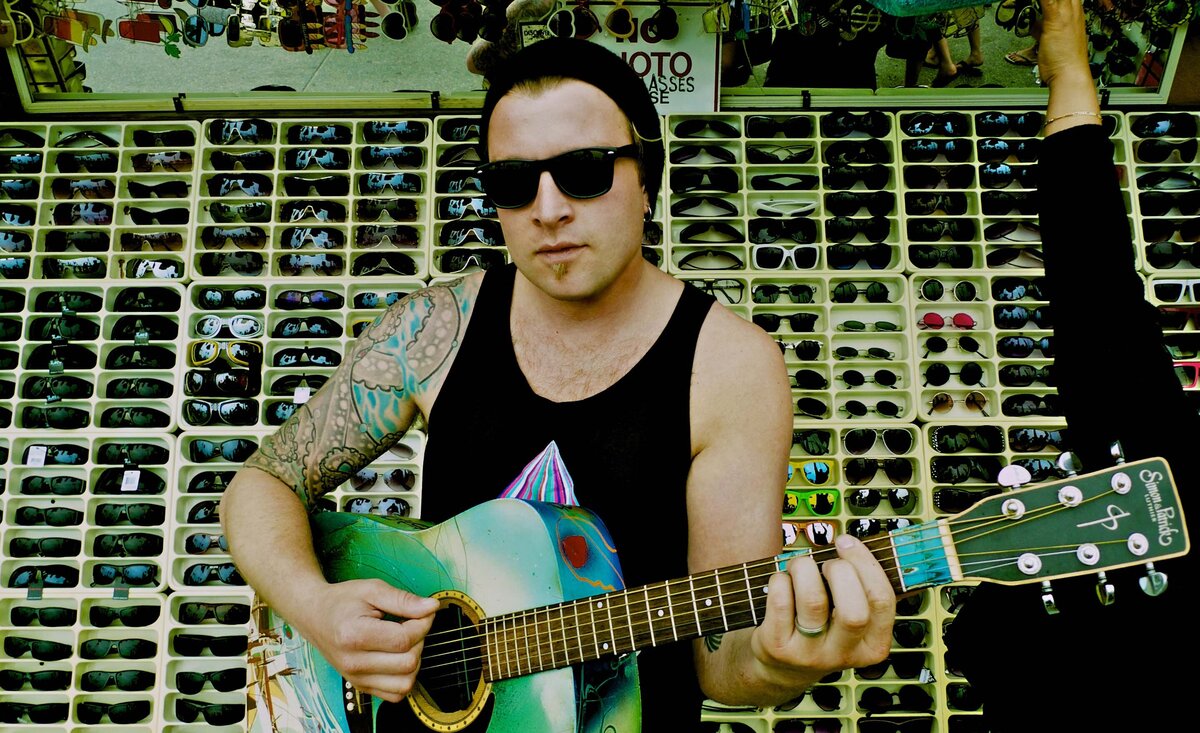 Male musician photo Daniel Wesley wearing black t shirt with sunglasses holding pale blue guitar sunglass stand behind