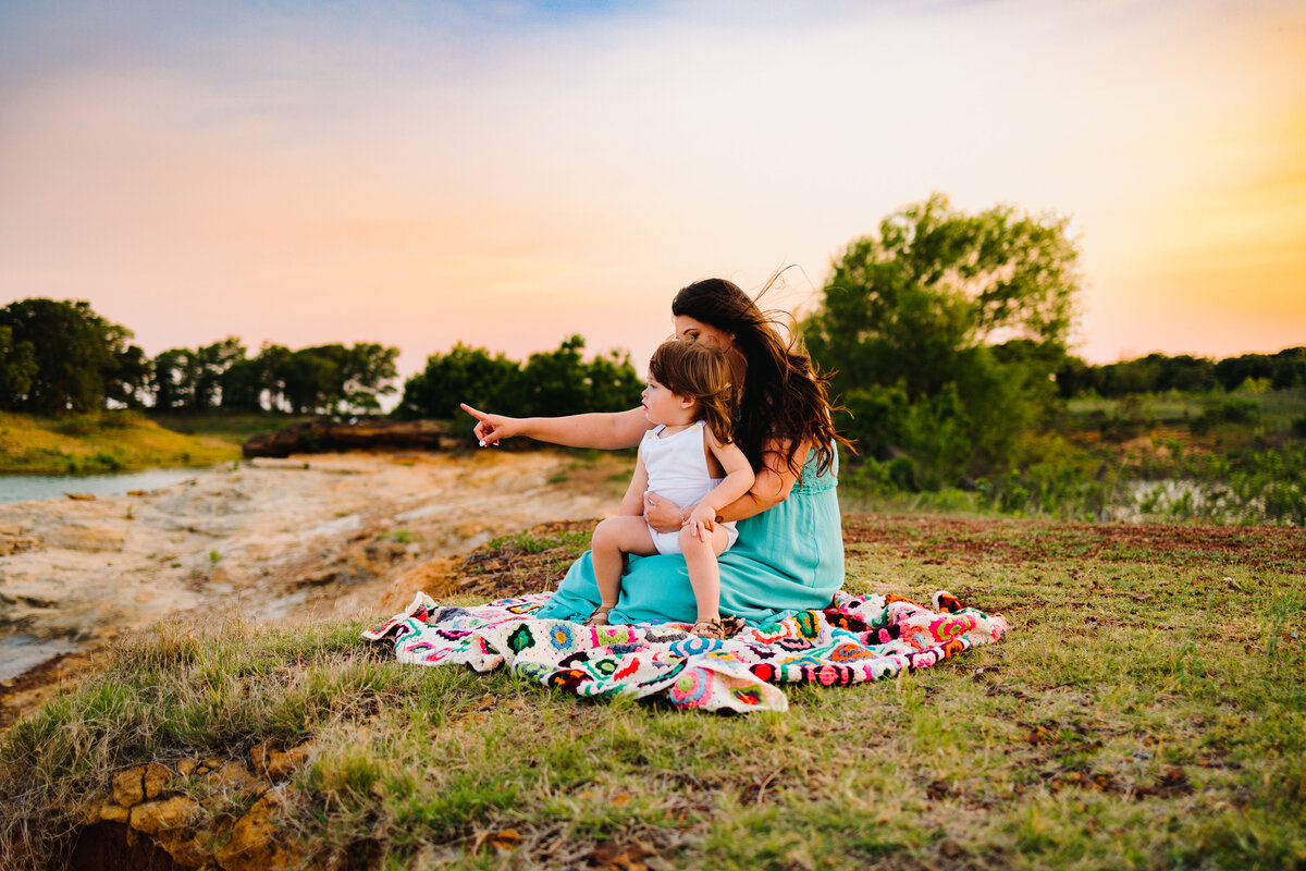 Photo of a mother with her daughter sitting on a colorful blanket. She is in an aquamarine dress and is pointing to the left side while carrying the girl in a white dress