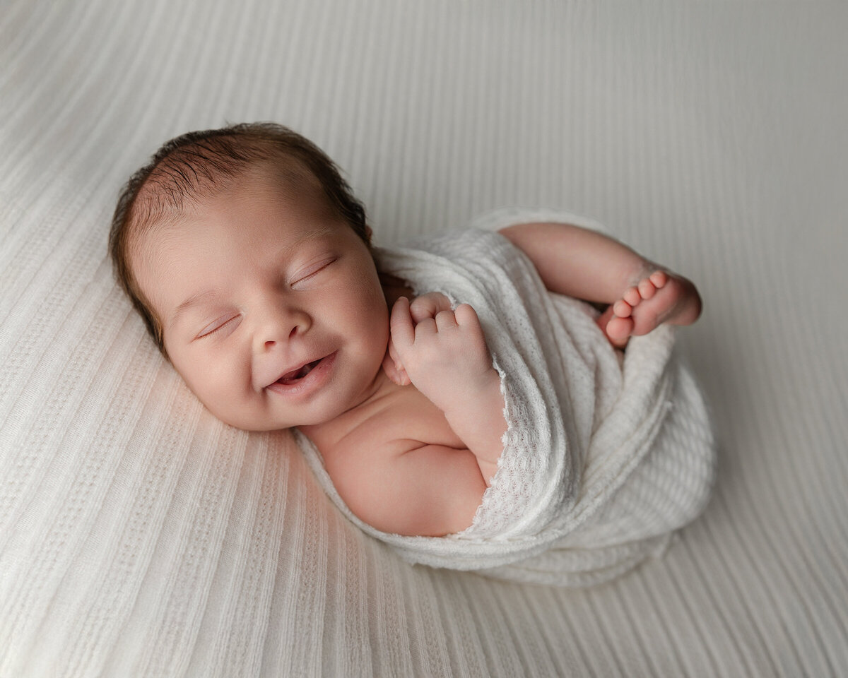 Newborn baby boy smiling in his sleep while swaddled in soft white textures, posed on a ribbed blanket during his studio session. Captured by Inland Empire newborn photographer Kaitlyn Dawn Photography.