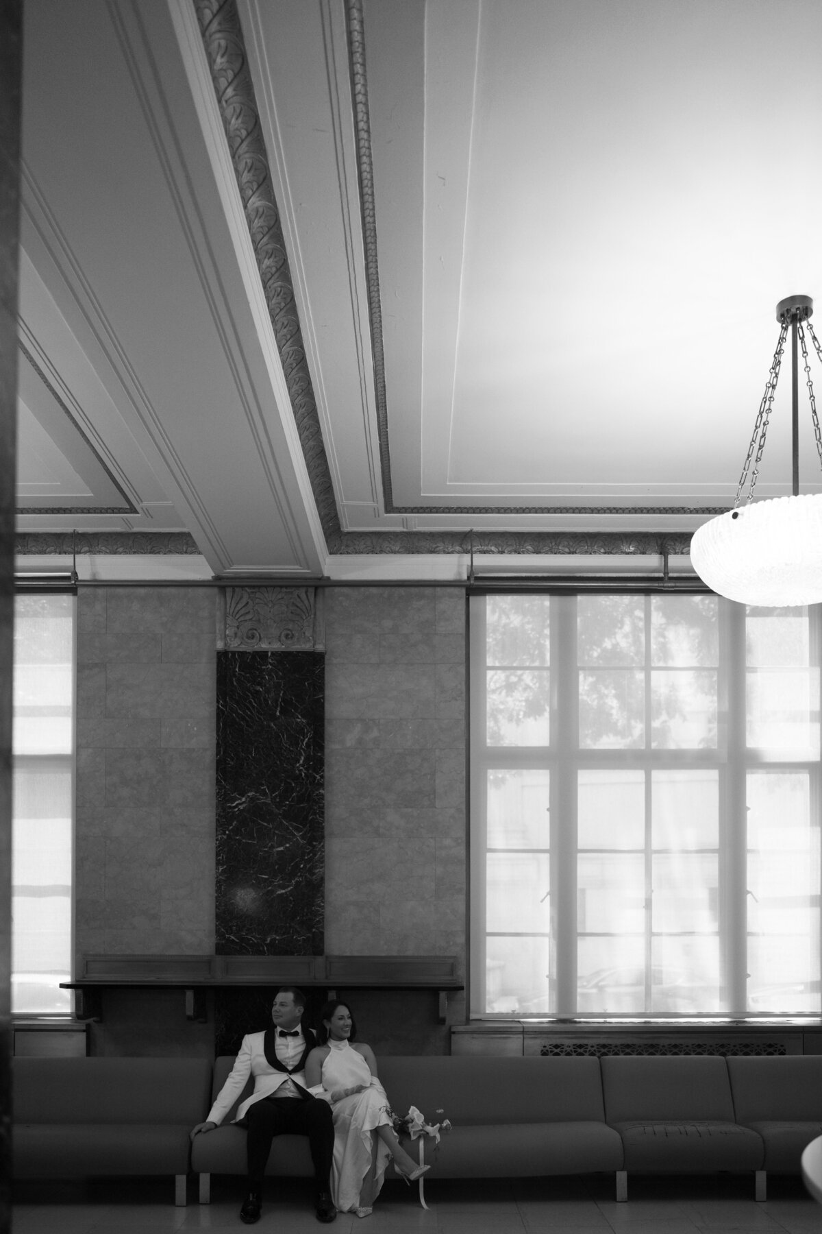 Black and white photo of the grand interior architecture inside New York City Hall, showing tall windows and chandeliers, captured during Japna and Chris’s elopement by NYC wedding photographer Perry Hancock.