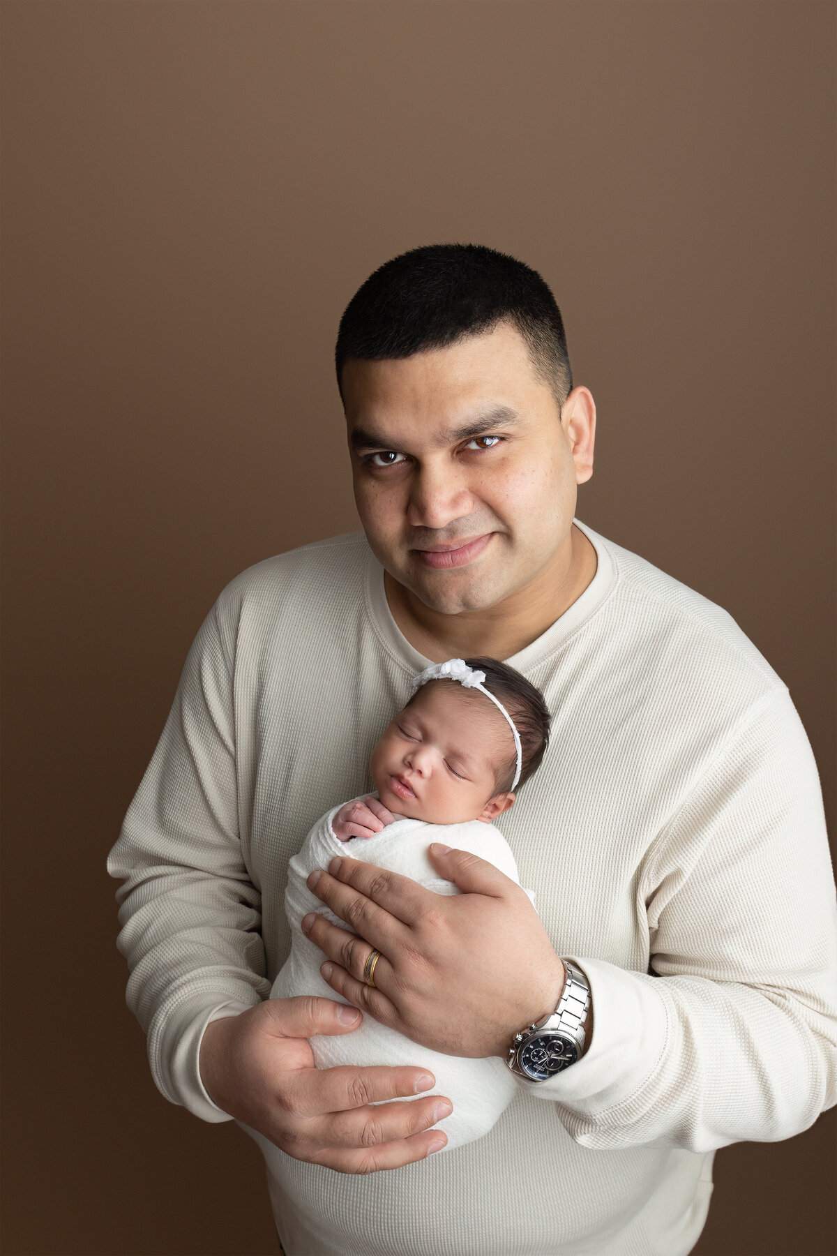 A tender studio portrait of a father cradling his newborn daughter in front of a warm brown backdrop. Photographed in my studio in Paris, Ontario.