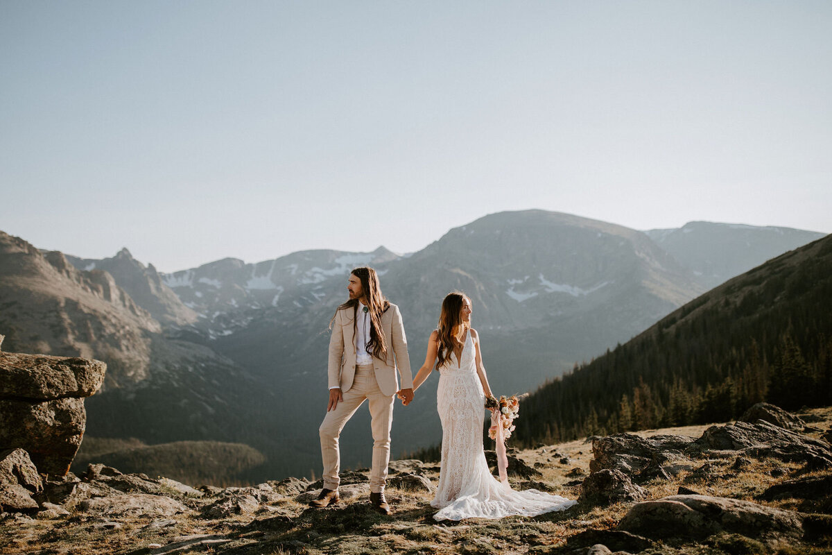 A bride and groom wearing an ivory suit and white wedding gown hold hands facing opposite directions on a mountain landscape.