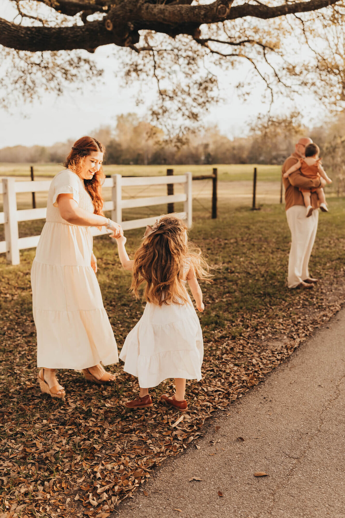 little girl dances with mom while dad holds daughter off to the side of the white picket fence.