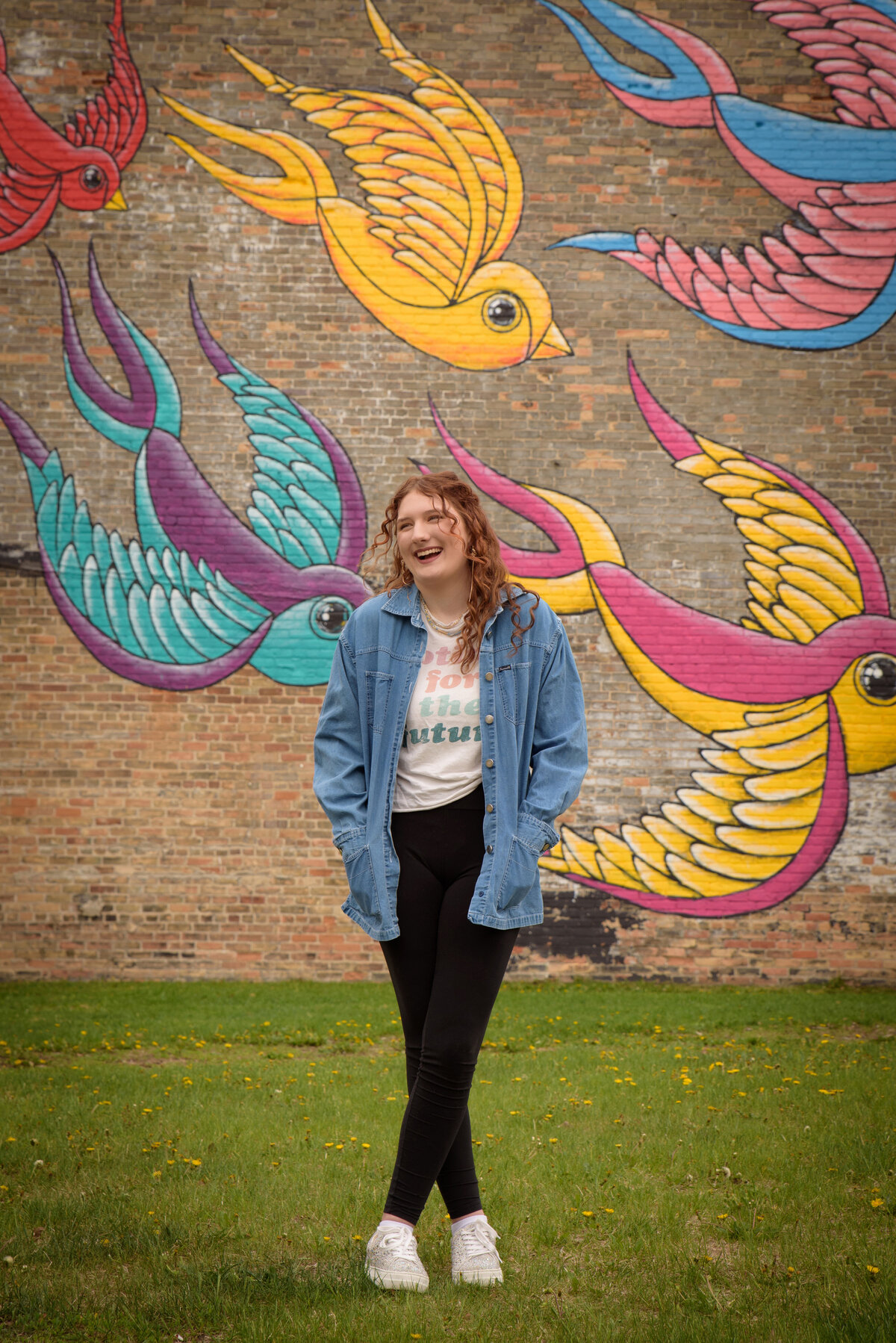 Green Bay East High School senior girl wearing a graphic t-shirt and leggings standing in front of a painted bird mural on a brick wall in an urban setting in downtown Green Bay, Wisconsin