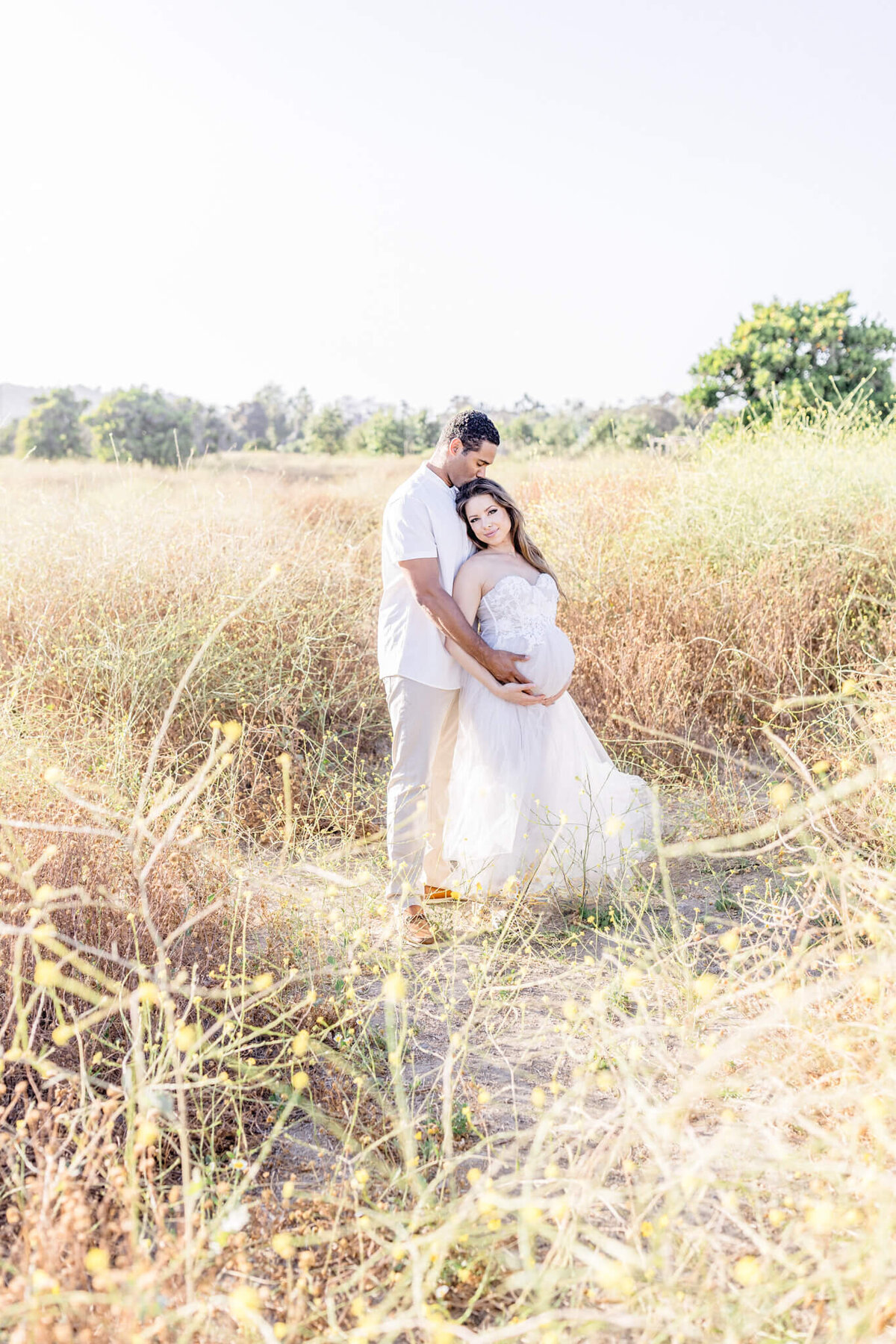A couple stands in a field of tall grass, embracing each other. The woman is wearing a flowing white dress, and the man is dressed in a white shirt and beige pants. The scene is bright and serene, with greenery in the background.