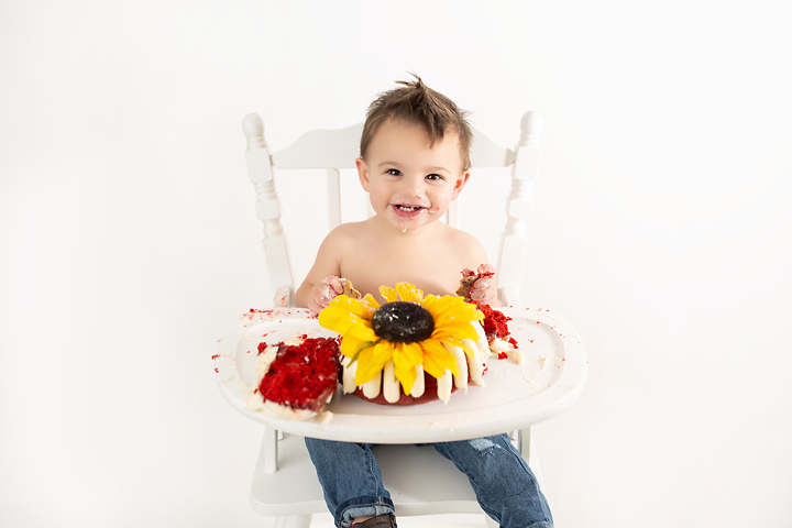 A toddler boy sits in a high chair with a sunflower designed red velvet cake