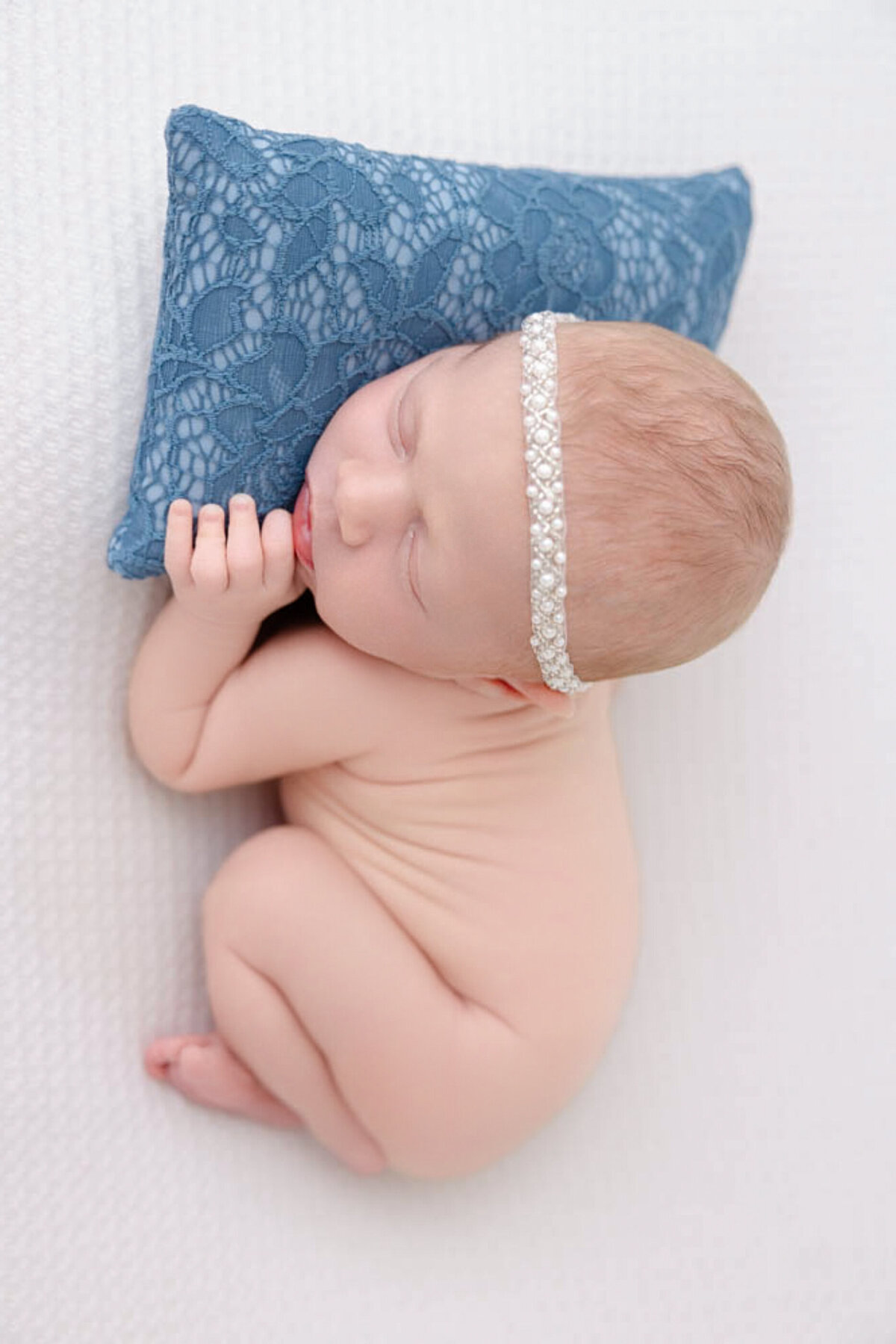 Baby girl wearing a pearl headband and lying on a blue laced pillow in bare skin.
