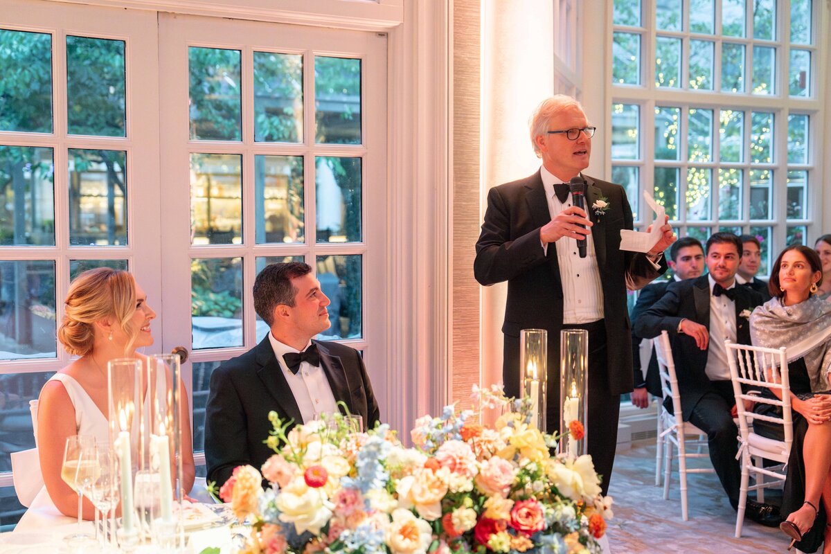 A man in a tuxedo speaks into a microphone at a formal event. A woman in a white dress and a man in a black tuxedo sit at a table with flowers, listening. Guests in formal attire are seated around them in a room with large windows.