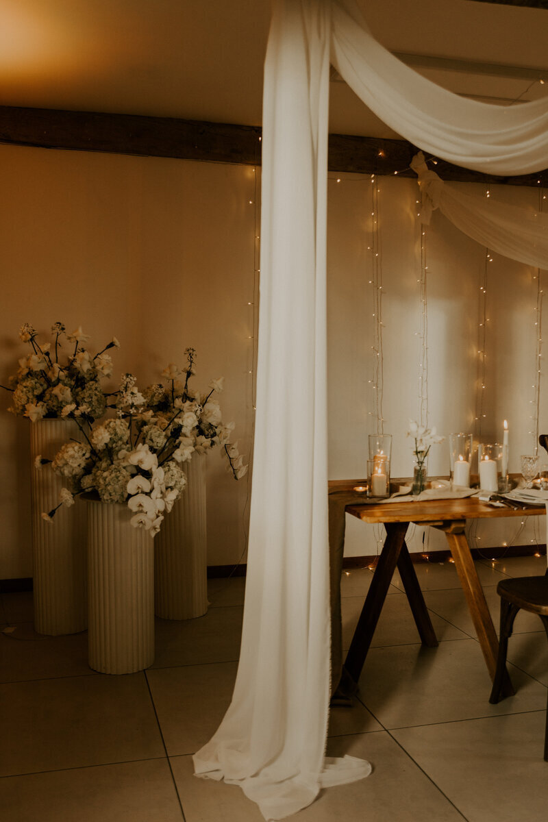 Table en bois encadrée d'un voilage et hauts pots surmontés de fleurs lors d'une séance photo mariage en vendée.