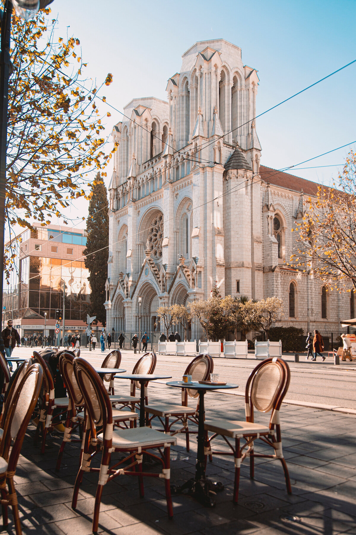 Street view of a European-style cathedral with ornate architecture in a sunny setting. A European travel specialist might highlight the cobblestone street in the foreground, where empty tables and chairs under leafless trees create an inviting atmosphere perfect for personalized travel planning.