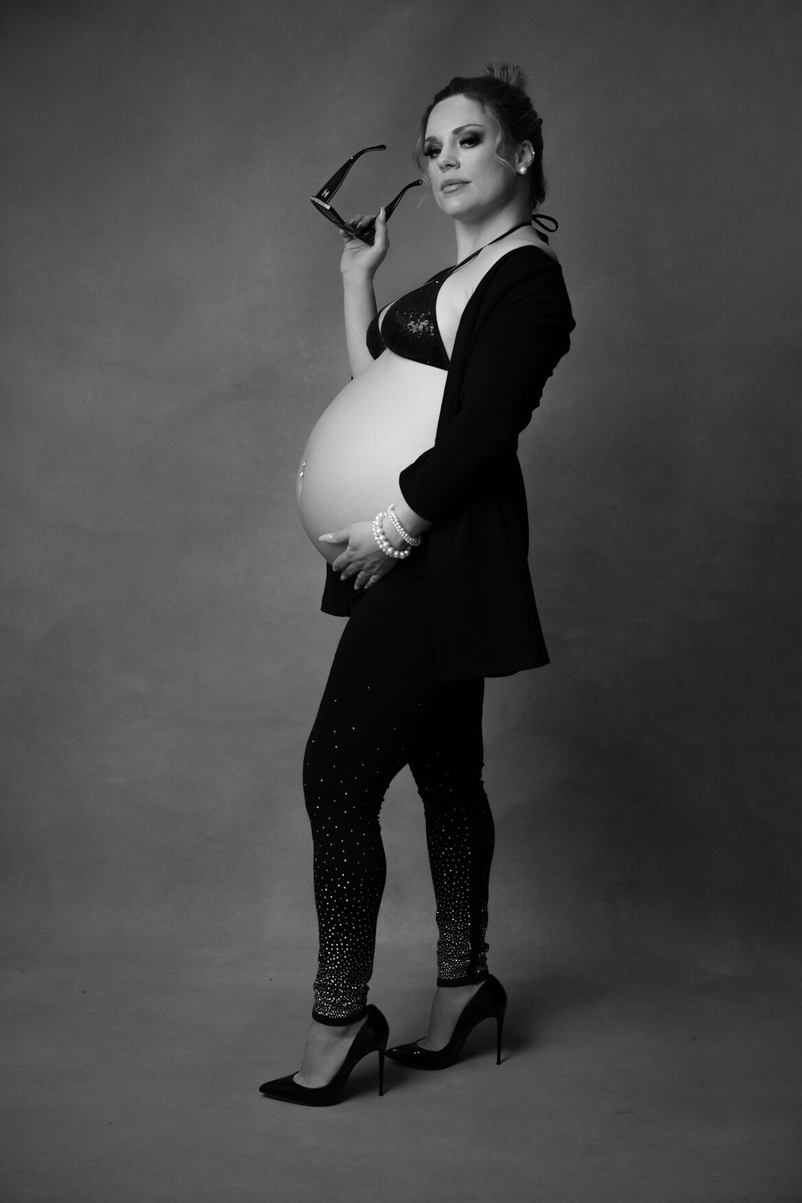 A pregnant woman posing confidently  in a dark background wearing black leggins and a black jacket holding sunglasses on her hand and with the other touching her naked belly