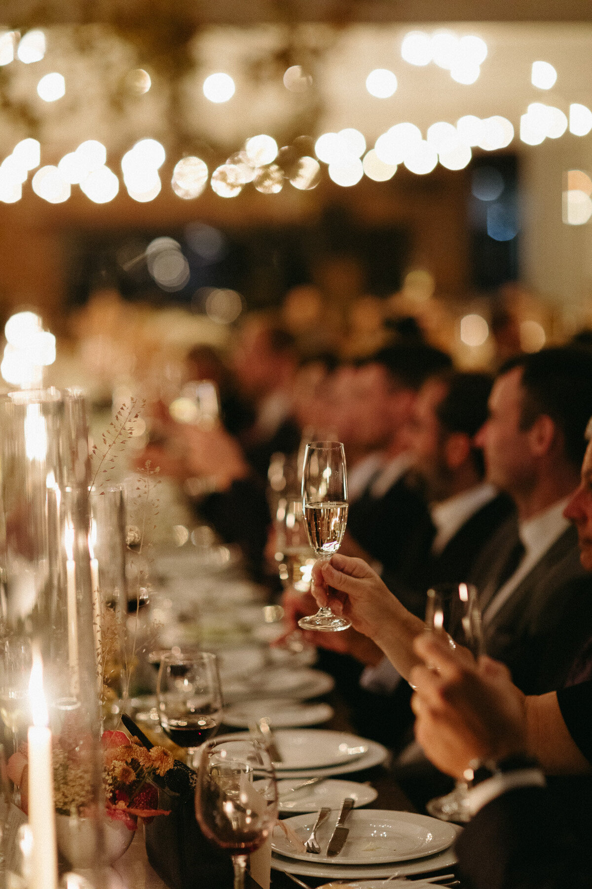 People seated at a long table raise glasses in a toast at a formal dinner event. Captured by an Oshawa wedding photographer, the candles and soft lighting create a warm atmosphere.