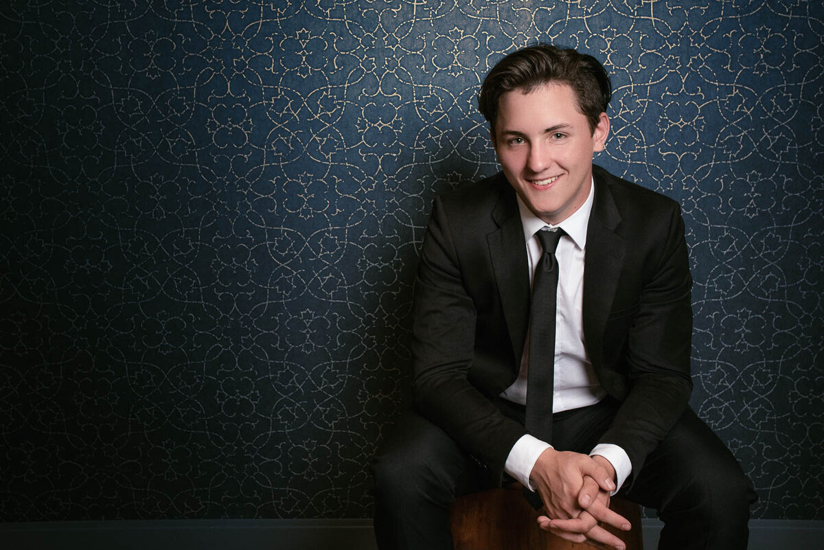Senior portrait of a teen boy in a black suit, smiling warmly while seated indoors in front of a blue and gold patterned wall.