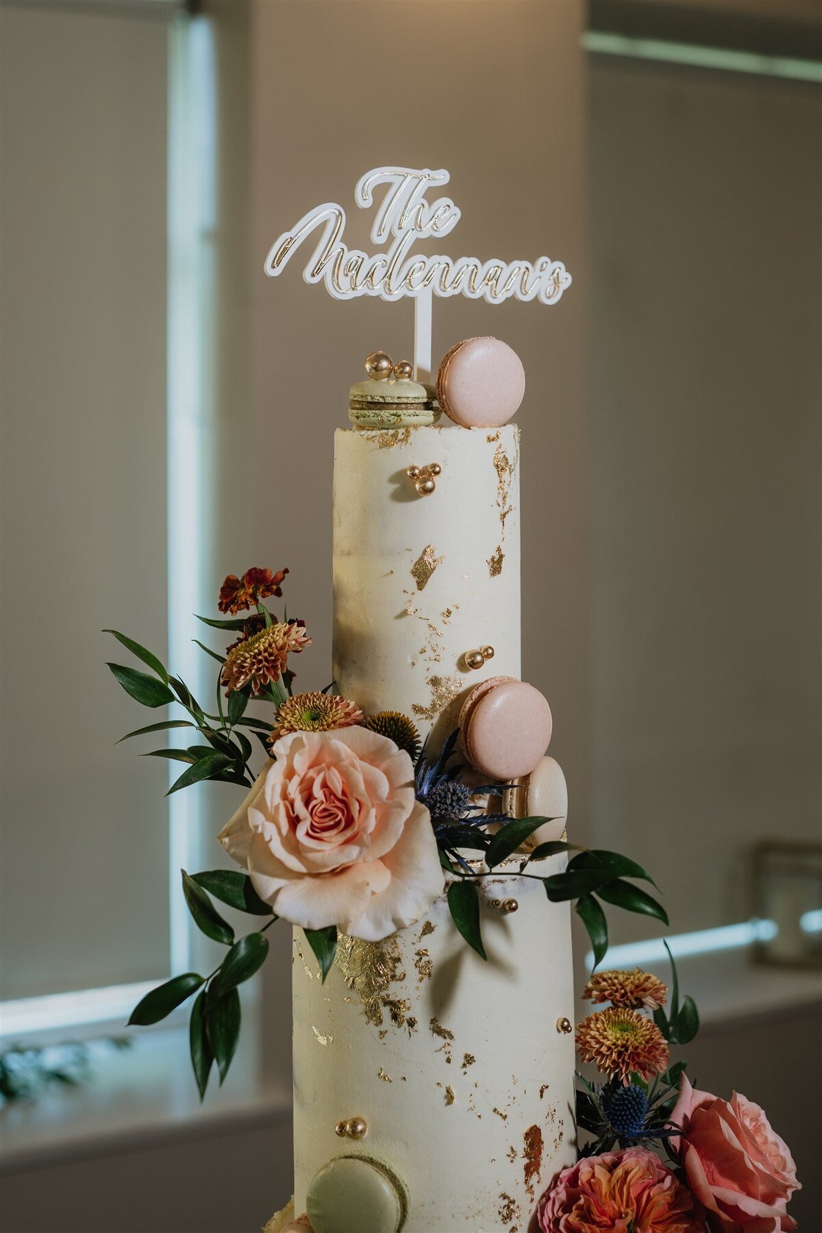 A three tier wedding cake stands on a rustic barrel. The cake is decorated with cream icing, roses, macarons, and foliage.