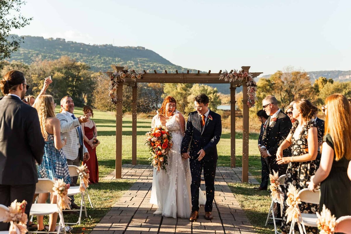 A newlywed couple walks hand in hand down the aisle, beaming as they exit their outdoor wedding ceremony beneath a wooden arbor adorned with dried florals in warm autumn tones. The bride, a red-haired woman in a flowing white gown with delicate lace details, carries a cascading bouquet of vibrant orange and red flowers. She laughs with joy as guests shower them with celebratory confetti, the golden sunlight catching the sparkles in the air. The groom, dressed in a navy plaid suit with an orange boutonniere, looks down with a soft smile as they take their first steps as a married couple. Guests, dressed in colorful attire, stand and cheer, tossing confetti with outstretched hands. The backdrop features rolling hills, lush greenery, and a clear blue sky, framing the couple in a perfect autumn wedding scene